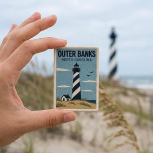 May include: A rectangular embroidered patch featuring a light blue sky, a black and white striped lighthouse, a small white house, and tan sand dunes. The text reads OUTER BANKS NORTH CAROLINA in dark blue letters against the sky background.