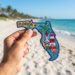 May include: An embroidered patch shaped like the state of Florida with a blue border. It features the text FLORIDA in yellow, a red star, a green palm tree, a white space shuttle, a red and white lighthouse, and a pink flamingo on blue waves.
