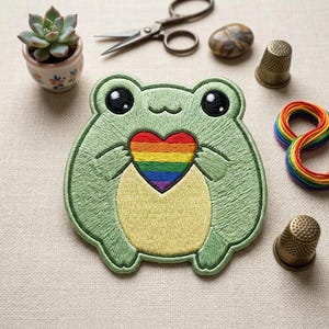 May include: An embroidered patch featuring a light green frog with a pale yellow belly, large black eyes, and a small smile, holding a heart with horizontal stripes in red, orange, yellow, green, blue, and purple colors against a light fabric.