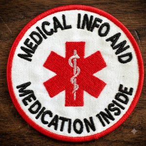 May include: A circular embroidered patch with a white background and a red border. It features a red Star of Life symbol in the center with a white caduceus. The black text reads MEDICAL INFO AND MEDICATION INSIDE in a circular arrangement.