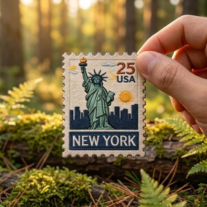 May include: A rectangular embroidered patch designed like a postage stamp featuring the Statue of Liberty in green, a dark blue city skyline, a yellow sun, and the text 25 USA and NEW YORK in dark blue and brown thread on a cream background.