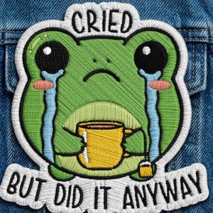 May include: An embroidered patch featuring a green frog with large black eyes, crying blue tears, and holding a yellow mug with a tea bag. The text above reads CRIED and the text below reads BUT DID IT ANYWAY, all outlined in white thread.