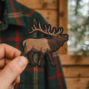 May include: An embroidered patch featuring a brown elk with large antlers in profile. The elk has a dark brown neck and head with a lighter tan body, all outlined in a thick black border. The design captures the animal in a standing position.