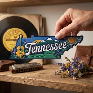 May include: An embroidered patch shaped like the state of Tennessee featuring a yellow acoustic guitar, purple iris flowers, green mountains, and a golden sun. The word Tennessee is written in white script across the center of the patch.