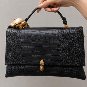May include: A black handbag with a textured crocodile-embossed pattern, featuring a single top handle and a gold-toned peanut-shaped clasp. A small, patterned silk scarf is tied to the base of the handle on the left side of the bag.
