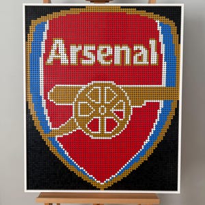 May include: A framed mosaic of the Arsenal Football Club emblem. The logo features a red shield with the word "Arsenal" in white, a cannon, and blue and gold detailing. The mosaic is constructed from small, colourful pieces.