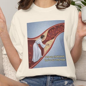 Op de afbeelding: Een off-white t-shirt met korte mouwen en een grafische print van een persoon in een wit gewaad in een bloedvat met rode bloedcellen en gele cholesterolophoping. De tekst luidt: Help us, cholesterol. You're our only hope.