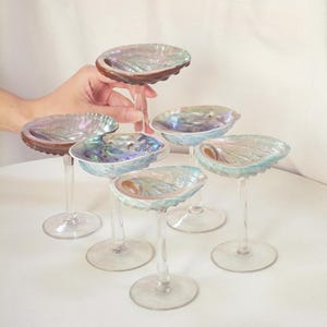 Könnte beinhalten: Ein Set aus fünf Stielgläsern mit Schalen aus irisierenden Abalone-Muscheln. Jedes Glas hat einen klaren, schlanken Stiel und einen runden Fuß. Die Muscheln weisen natürliche, wirbelnde Muster in Blau-, Grün- und Violetttönen mit glänzendem Finish auf.