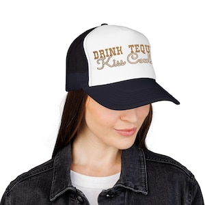 May include: A black and white trucker hat featuring a white front panel with the brown embroidered text "DRINK TEQUILA Kiss Cowboys". The hat has a black brim and a black mesh back panel, designed with a structured front and an adjustable closure.