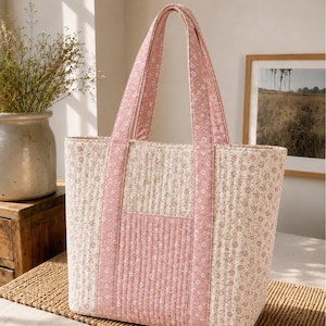 Può includere: Una borsa tote trapuntata in tessuto color crema con un piccolo motivo floreale rosa, arricchita da pannelli verticali trapuntati in rosa tinta unita e manici abbinati. La borsa ha una forma rettangolare e una consistenza morbida e cucita.