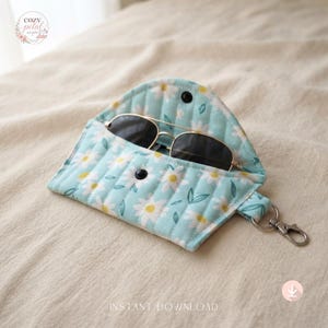 May include: A quilted fabric glasses case in light blue with a white daisy floral pattern, featuring a snap button closure and a metal lobster clasp keychain attachment. The case is open and contains a pair of dark-tinted aviator sunglasses.