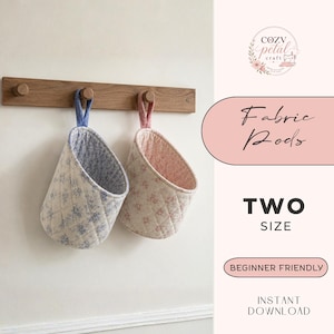 Op de afbeelding: Twee gewatteerde stoffen mandjes met bloemenpatronen, één in blauw en één in roze, elk voorzien van een stoffen ophanglus. De mandjes hangen aan een houten wandrek met haken. De tekst op de afbeelding luidt Fabric Pods, TWO SIZE, BEGINNER FRIENDLY, INSTANT DOWNLOAD en COZY petal CRAFT.