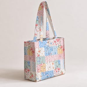 May include: A quilted tote bag featuring a patchwork design of various floral and gingham patterns in soft shades of pink, blue, yellow, and green. The bag has two matching fabric handles and a rectangular structure with vertical stitching.