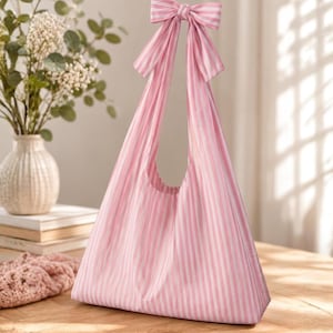 May include: A pink and white vertically striped fabric tote bag featuring a large bow detail at the top of the handle. The bag has a triangular silhouette with a wide base and a single integrated strap that forms the bow closure at the top center.