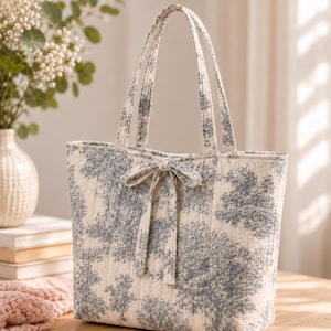 Puede incluir: Un bolso tote acolchado con un estampado toile de jouy azul y blanco de árboles y follaje. El bolso tiene dos asas verticales y un lazo decorativo de tela atado en el centro de la abertura superior. La textura acolchada es visible en todo el diseño.