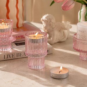 Pink glass tealight candle holders styled on a neutral stone tabletop with books, tulips, and soft editorial lighting.