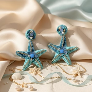 Op de afbeelding: Een paar turquoise en blauwe, met kralen bezette zeesteroorbellen. De oorbellen hebben een vijfhoekige stervorm, gedetailleerd met kralenwerk, parels en blauwe edelstenen. Ze hangen aan een cirkelvormige oorbel en zijn bevestigd aan een gouden ketting.
