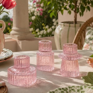 May include: Three pink glass candle holders with a ribbed texture sit on a table. The candle holders vary in height and design, with a fluted edge on the tallest. A vase of tulips and a plate are in the background.