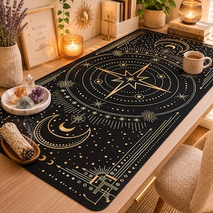 May include: Black desk mat with a gold celestial design featuring a compass rose, stars, and moons. The mat is rectangular and sits on a wooden desk. Other items on the desk include candles, crystals, and a framed print that says "BE STILL AND KNOW".