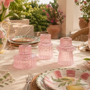 Pastel pink glass candle holders arranged as centrepieces on a cream linen outdoor dining table with colourful plates and dopamine decor styling.
