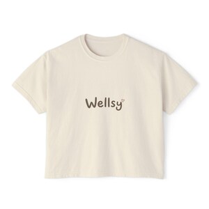 Puede incluir: Camiseta color crema con la palabra "Wellsy" en escritura marrón, un pequeño corazón rosa está al lado de la "y". La camiseta tiene cuello redondo y mangas cortas. La tela parece ser de un material de algodón suave.