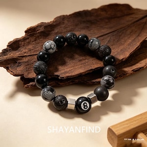 Könnte beinhalten: Ein Perlenarmband aus einer Mischung von schwarzen und grauen Steinperlen, darunter texturiertes Lavagestein, glatter Obsidian und Schneeflocken-Obsidian, akzentuiert durch zwei silberfarbene Metallwürfel und eine zentrale schwarze Perle mit einer weißen Acht.