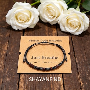 Könnte beinhalten: Ein schwarzes Schnurarmband mit abwechselnden dunkelbraunen Holzperlen und kleinen metallisch grauen Perlen in einem Morsecode-Muster. Das Armband liegt auf einer beigen Karte mit der Aufschrift Morse Code Bracelet und Just Breathe.