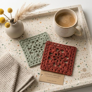 May include: Two square crochet coasters in sage green and terracotta orange with a floral motif, a beige ceramic mug with coffee, a small vase with yellow billy balls and pampas grass, and a beige waffle-weave cloth on a speckled tray.