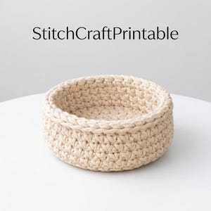 May include: A round, beige, crocheted basket with a thick, textured stitch pattern and a reinforced rim. The text StitchCraftPrintable is displayed in black above the basket, which features a consistent, neutral color throughout its design.