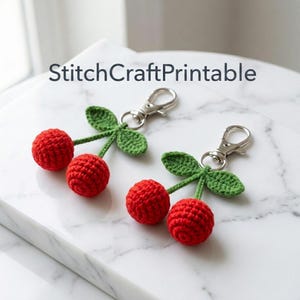 Op de afbeelding: Twee gehaakte kersensleutelhangers met felrode bolvormige vruchten en groene blaadjes, bevestigd aan zilverkleurige metalen karabijnsluitingen. De tekst StitchCraftPrintable is erboven zichtbaar. Elk kersenpaar is voorzien van een steeltje en twee blaadjes.