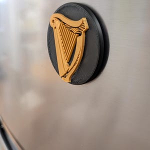 May include: A black and gold 3D-printed magnet featuring the Guinness harp logo. The harp is gold, set against a black background, and mounted on a silver surface. The magnet is round and has a textured appearance.