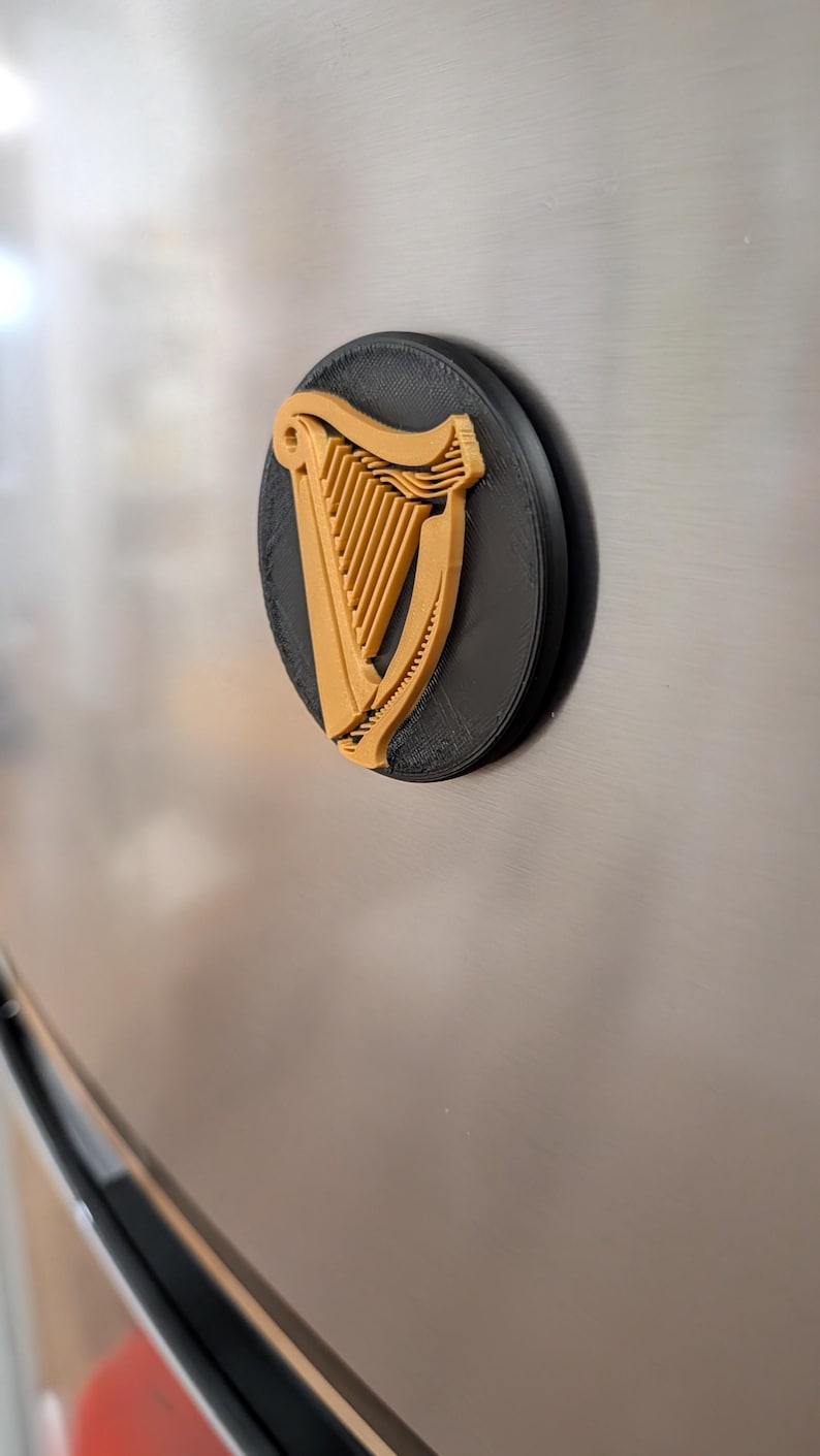 May include: A black and gold 3D-printed magnet featuring the Guinness harp logo. The harp is gold, set against a black background, and mounted on a silver surface. The magnet is round and has a textured appearance.