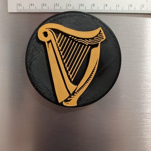 May include: A round, black magnet featuring a gold-colored harp design. The magnet is approximately 3 inches in diameter and is placed on a metallic surface. The harp is a prominent symbol.