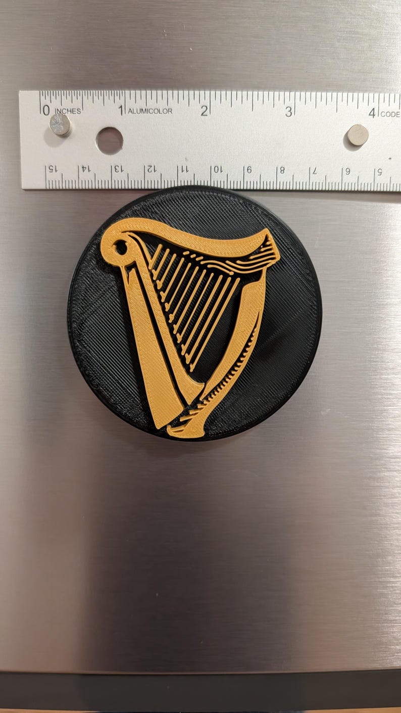 May include: A round, black magnet featuring a gold-colored harp design. The magnet is approximately 3 inches in diameter and is placed on a metallic surface. The harp is a prominent symbol.