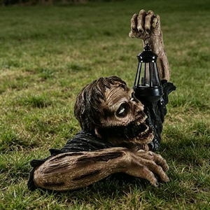 Könnte beinhalten: Eine Halloween-Gartendekoration, die einen Zombie zeigt, der aus dem Boden auftaucht. Der Zombie hat ein verfallenes Gesicht und ausgestreckte Arme, die eine schwarze Laterne halten. Die Dekoration befindet sich in grünem Gras.