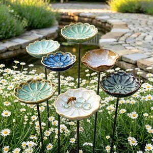 Könnte beinhalten: Eine Sammlung von keramischen, blütenförmigen Vogeltränken in verschiedenen Farben, darunter Blau, Grün und Creme. Jede Tränke ist auf einem schwarzen Metallstab montiert, der in einem Garten mit Gänseblümchen steht.