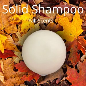 May include: A solid, round shampoo bar in a cream color, resting on a bed of colorful autumn leaves. The text "Solid Shampoo" and "Fall Scents" are displayed in white font above the bar, suggesting a seasonal fragrance.