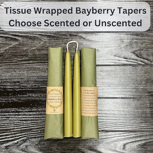 May include: Two olive green taper candles connected by a wick, partially wrapped in green tissue paper with tan labels. The labels feature text including Hand Dipped Unscented Real Bayberry Wax Taper Candles and The Bayberry Candle Legend.