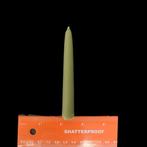 May include: A single, tall, green candle measuring approximately 7.6 cm in length. The candle is positioned on top of a red ruler with black markings.