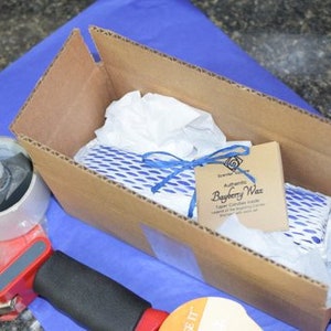 May include: A cardboard box with a blue and white patterned item inside. The box is labeled "Authentic Bayberry Wax" and has a blue ribbon tied around it.