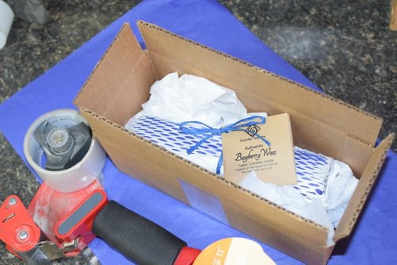 May include: A cardboard box with a blue and white patterned item inside. The box is labeled "Authentic Bayberry Wax" and has a blue ribbon tied around it.