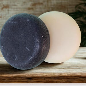May include: Two round soaps, one dark gray and the other a creamy off-white, sit side-by-side on a wooden surface. The dark gray soap has a speckled appearance, while the off-white soap has a smooth texture. The soaps are likely for body care.
