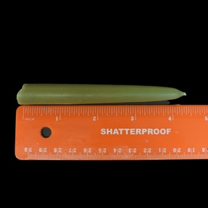 May include: A green candle measuring approximately 10 cm in length. The candle is lying on a ruler with the text "SHATTERPROOF" printed on it.