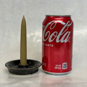 May include: A small, black ceramic candle holder with a single, tall, beige candle. The candle holder is next to a can of Coca-Cola.