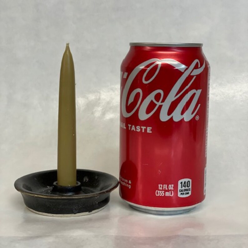 May include: A small, black ceramic candle holder with a single, tall, beige candle. The candle holder is next to a can of Coca-Cola.