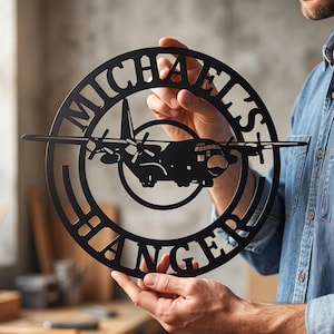 May include: A circular black metal wall sign featuring a silhouette of a four-engine propeller aircraft in the center. The text MICHAELS is cut out along the top inner edge of the circle, and the word HANGER is cut out along the bottom edge.