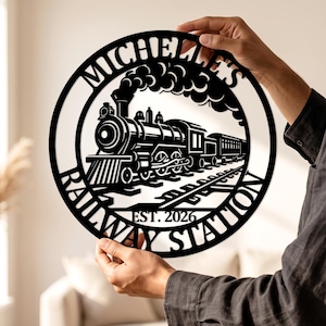 May include: A circular black metal wall sign featuring a cutout design of a steam locomotive train on tracks with smoke billowing from the chimney. The text MICHELLE'S RAILWAY STATION is arched around the border, with EST. 2026 at the bottom.