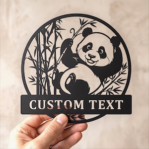 Peut inclure: Une enseigne circulaire en métal noir découpé représentant un panda assis parmi des tiges de bambou. Le design comprend une barre rectangulaire horizontale en bas avec les mots CUSTOM TEXT en police serif, formant une décoration murale.