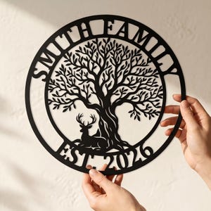 Könnte beinhalten: Ein kreisförmiges Wandbild aus schwarzem Metall mit einem lasergeschnittenen Design eines großen Baumes, an dessen Fuß ein Hirsch ruht. Der Schriftzug SMITH FAMILY ist oben gebogen und der Text EST. 2026 befindet sich am unteren Rand des Kreises.