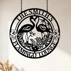 Peut inclure: Une enseigne murale circulaire en métal noir avec un motif découpé représentant deux flamants roses face à face, entourés de fleurs tropicales, de feuilles et d'ananas. Le texte indique THE SMITH'S et FLAMINGO LOUNGE. Suspendue par deux chaînes.